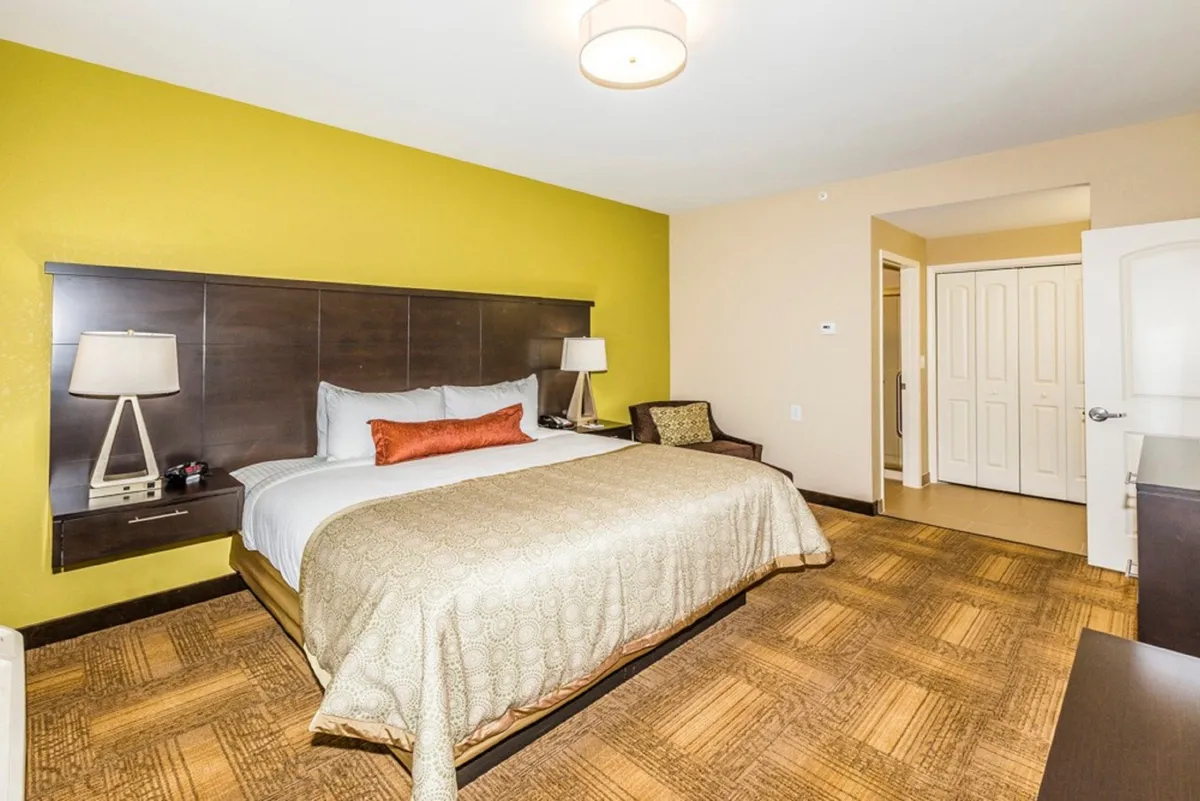 A spacious bedroom featuring a plush King bed adorned with crisp white linens, accented by decorative pillows and a cozy nightstand with a lamp. The soft lighting and gentle morning light filtering through the window create a comforting, restful atmosphere, ideal for relaxation and rejuvenation.