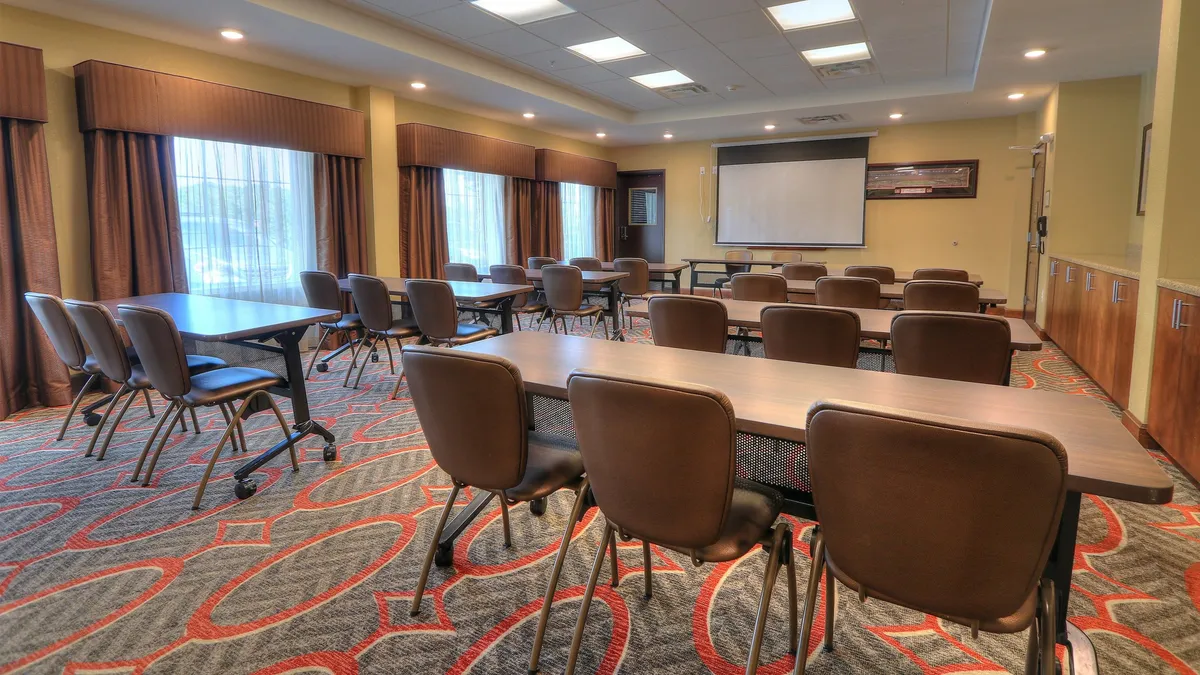 This image features a well-appointed meeting space with tables arranged for collaboration, comfortable chairs, and large windows letting in ample natural light. There's a prominent presentation screen in the background, creating an atmosphere conducive to business meetings, workshops, and networking events. The space feels modern and inviting, supporting productive professional activities.