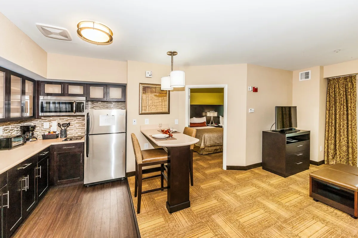 An open-plan interior with a view of the kitchen, a cozy living area with a pull-out sofa, a dining table, a TV unit, and the door to the bedroom. The space appears airy and welcoming, designed for lounging, dining, or unwinding with cable TV or a movie.