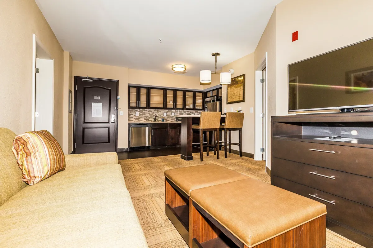 The image showcases a bright, open-plan living area with a plush sofa, a sleek dining table set for two, a large flat-screen TV, and a modern kitchen area. The cozy atmosphere is enhanced by warm lighting and inviting decor, perfect for relaxing, sharing meals, or unwinding after exploring Knoxville.