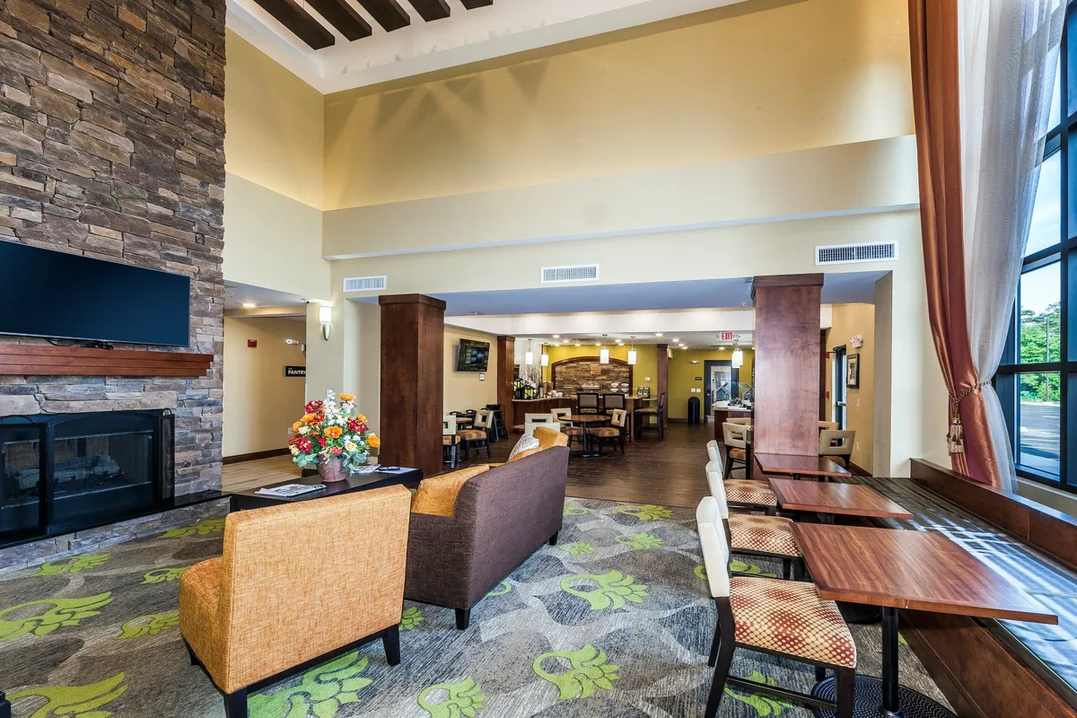 A warm, welcoming lounge area with a stone fireplace as its centerpiece, surrounded by a comfortable seating arrangement and large windows that flood the room with natural light. A decorative flower arrangement adds a touch of homeliness, and the serene atmosphere is ideal for reading, working, or unwinding.