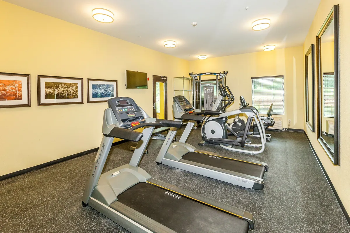 A well-equipped fitness center with treadmills, an elliptical machine, a TV screen for entertainment, large mirrors, and framed artwork. The overall feel is motivational and health-focused, supporting active lifestyles.