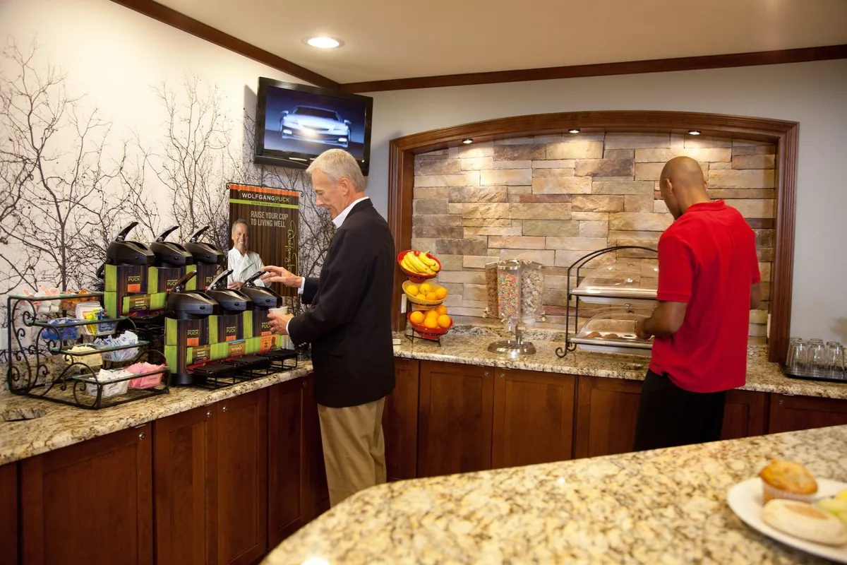 The image highlights a coffee station, a vibrant display of fresh fruits, and a relaxed communal atmosphere, with a stone accent wall and a television screen adding to the space's appeal. Guests are seen enjoying coffee and light breakfast, fostering a social and inviting ambiance.