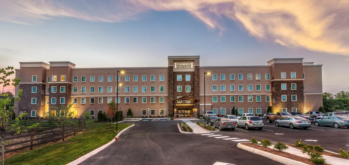 A welcoming view of the hotel entrance framed by lush landscaping and a spacious parking lot. The image captures families and small groups arriving with shopping bags, hinting at nearby retail opportunities. The golden hour lighting creates a warm, inviting mood.
